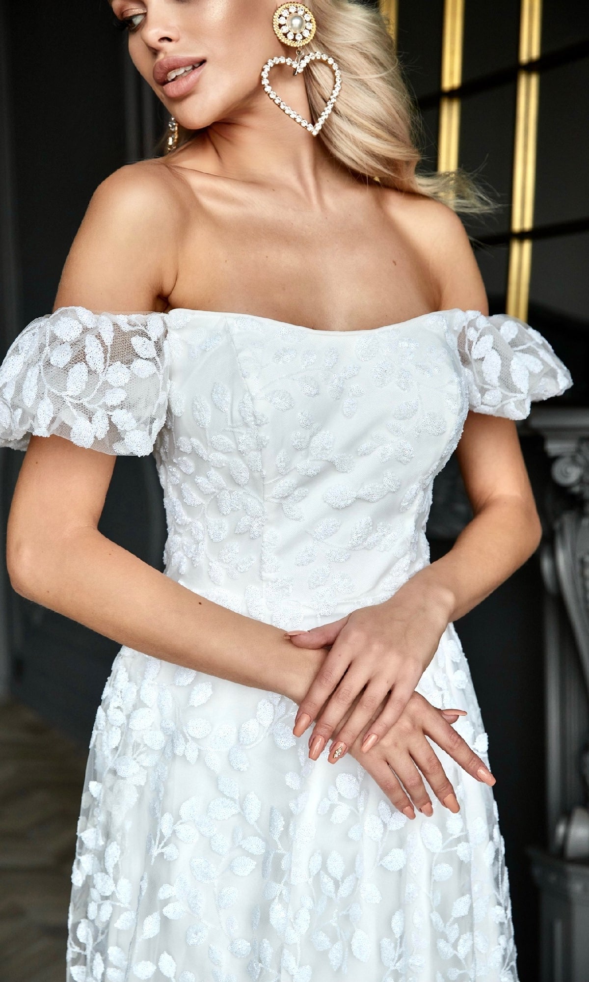 A woman stands indoors with arms crossed, wearing the Long White Lace Formal Dress: Tina Holly TK023W, featuring an off-the-shoulder design and sheer sleeves, accessorized with large heart-shaped pearl earrings.